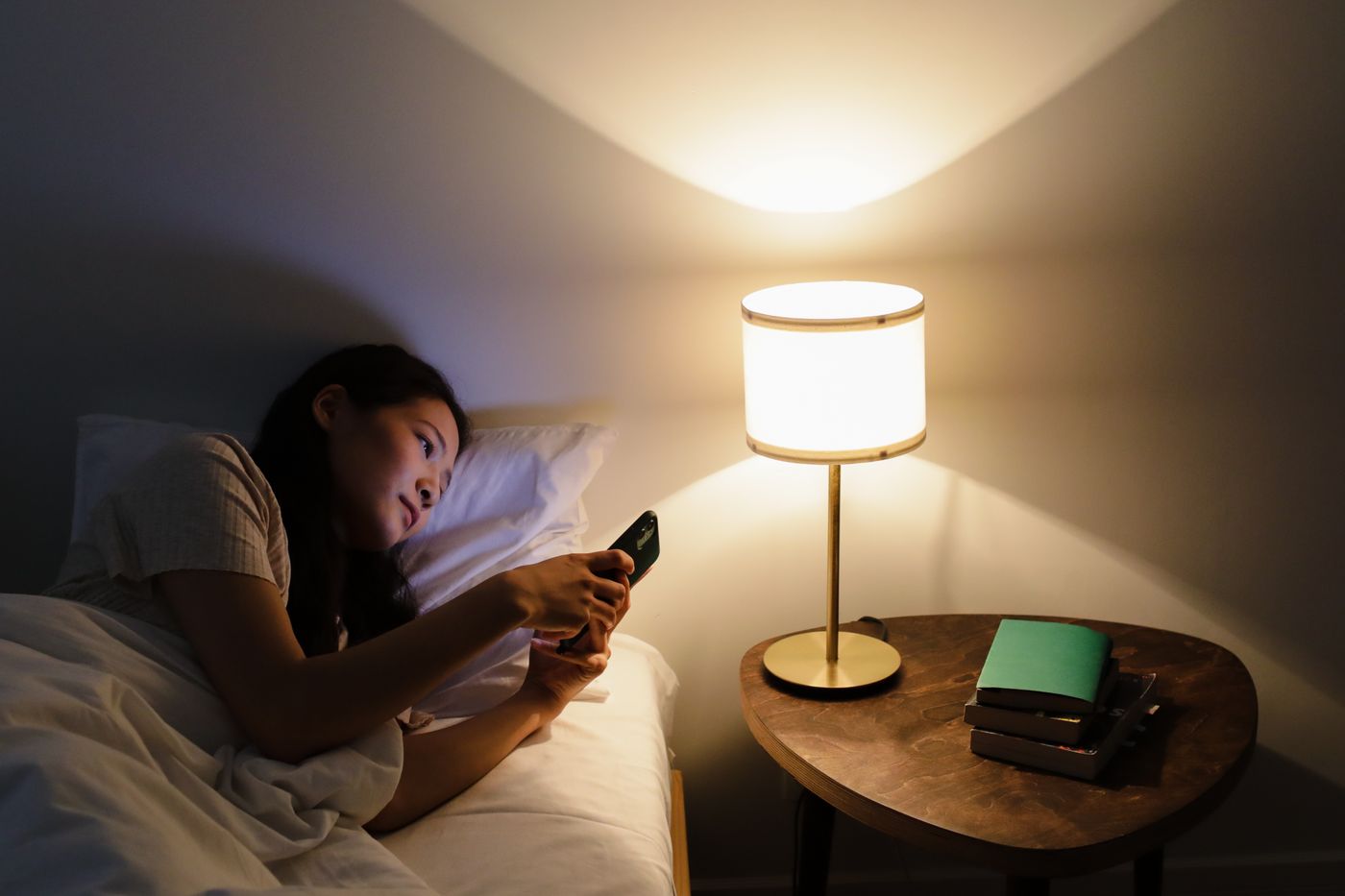 Frau liegt abends im Bett und bucht im Schein der Nachttischlampe auf dem Handy
